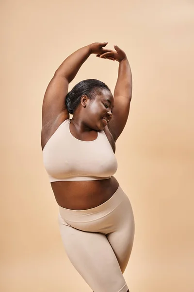 Happy plus size model in beige underwear posing with raised hands, body positive and self esteem — Stock Photo