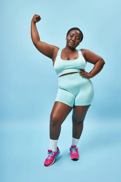 Energetic plus size woman in active wear flexing her muscles on blue background, hand on hip — Stock Photo
