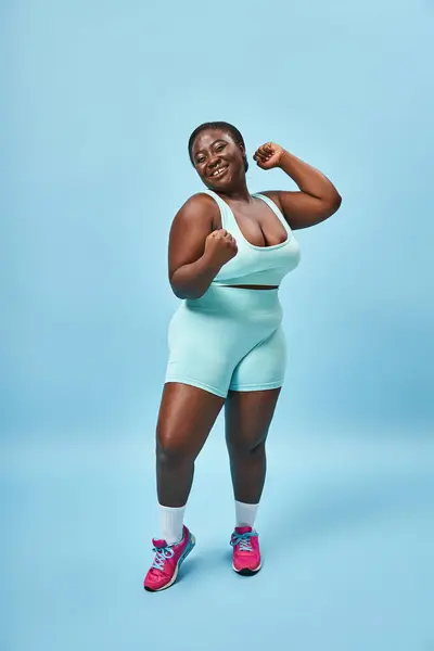 Excited plus size woman in active wear flexing muscles and looking at camera on blue background — Stock Photo