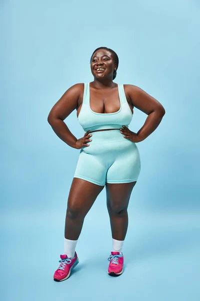 Felice plus size donna africana americana in blu abbigliamento sportivo con le mani sui fianchi sullo sfondo corrispondente — Foto stock