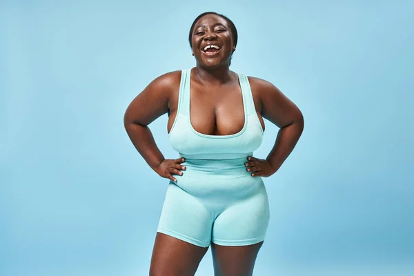 Happy plus size african american woman in blue sportswear with hands on hips on matching backdrop — Stock Photo