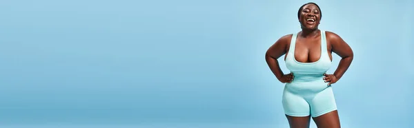 Счастливая плюс размер африканская американка в синей спортивной одежде позирует с руками на бедрах, баннер — стоковое фото