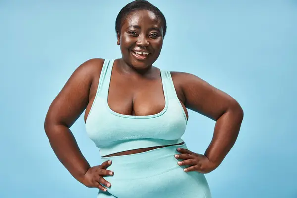 Heureux plus la taille femme afro-américaine en bleu vêtements de sport avec les mains sur les hanches sur fond assorti — Photo de stock