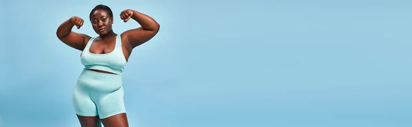 Banner, Plus Size Frau in aktiver Kleidung lässt ihre Muskeln spielen und schaut in die Kamera auf blauem Hintergrund — Stockfoto