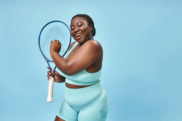 Smiling plus size african american woman ready to play tennis on blue background, body positive — Stock Photo