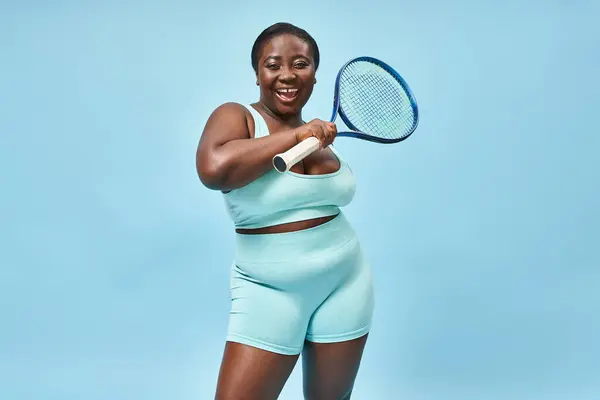 Alegre plus size mulher de pele escura segurando raquete de tênis no fundo azul, corpo esporte positivo — Fotografia de Stock