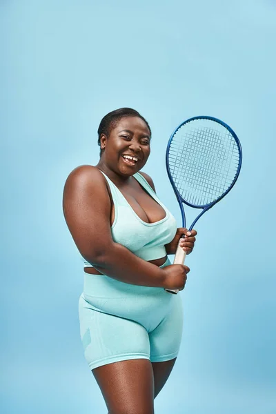 Feliz más tamaño negro mujer sosteniendo raqueta de tenis sobre fondo azul, cuerpo positivo y deporte - foto de stock