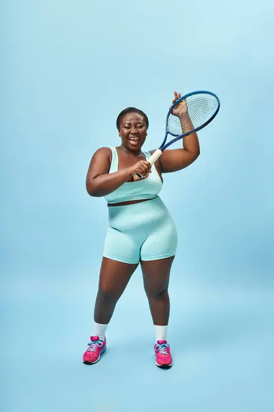 Excited plus size dark skinned sportswoman holding tennis racket on blue backdrop, body positive — Stock Photo