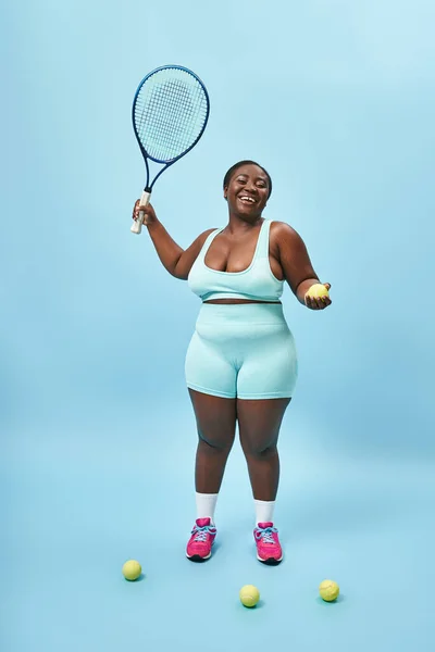 Animado plus size escuro pele esportista segurando raquete de tênis e bola no fundo azul, jogador — Fotografia de Stock
