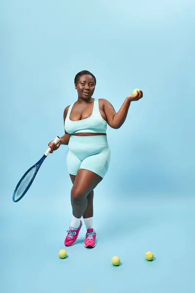 Feliz plus size esportista de pele escura segurando raquete de tênis e bola no fundo azul, jogador — Fotografia de Stock
