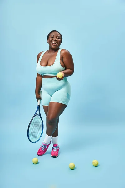 Cheerful plus size dark skinned sportswoman holding tennis racket and ball on blue backdrop, player — Stock Photo