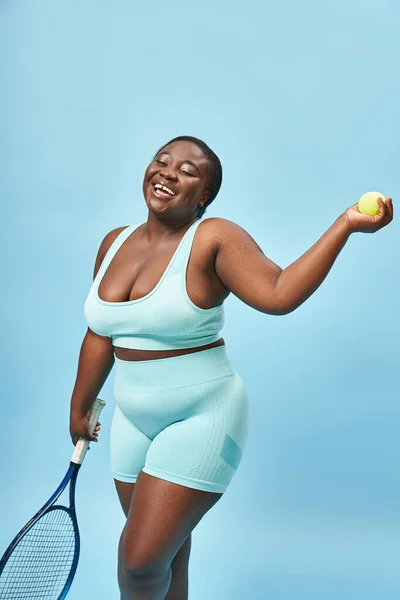 Sonriente más tamaño de piel oscura deportista sosteniendo raqueta de tenis y pelota sobre fondo azul, jugador - foto de stock