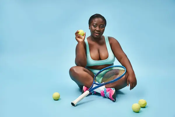 Молодые плюс размер темно-кожистая спортсменка держа теннисную ракетку и сидя вокруг шарики на синий — стоковое фото
