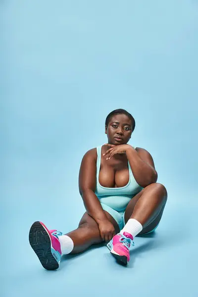 Relaxed plus size african american sportswoman sitting in blue sportswear on matching backdrop — Stock Photo