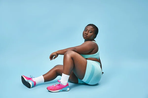 Relaxed plus size african american sportswoman sitting in blue sportswear on matching background — Stock Photo