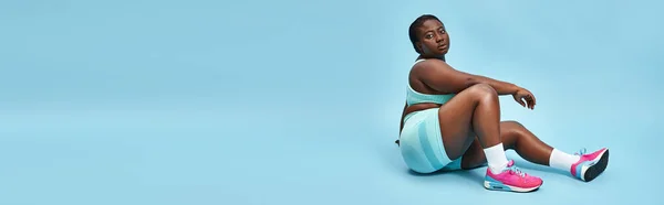 Relaxed plus size african american woman sitting in blue sportswear on matching backdrop, banner — Stock Photo