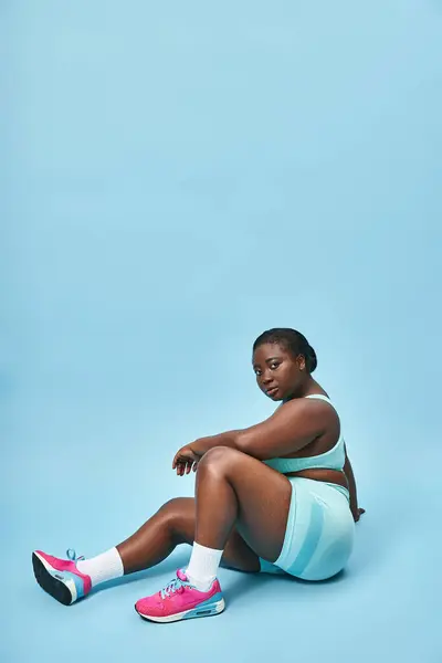 Relaxed plus size african american woman sitting in blue sportswear on matching backdrop, sport — Stock Photo