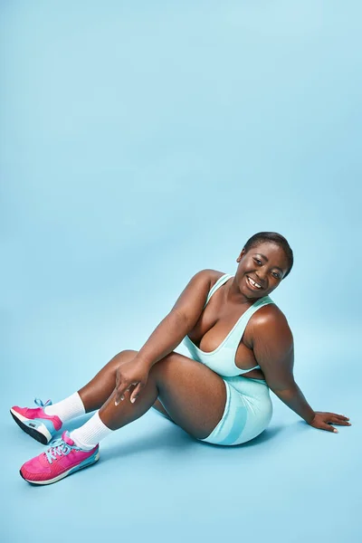 Счастливый плюс размер африканская американка сидит в синей спортивной одежде на соответствующем фоне, спорт — стоковое фото