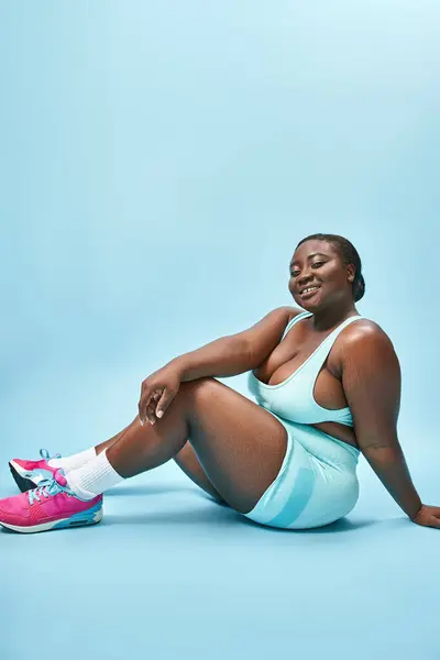 Sonriente más tamaño mujer afroamericana sentado en ropa deportiva azul en el telón de fondo a juego, deporte - foto de stock