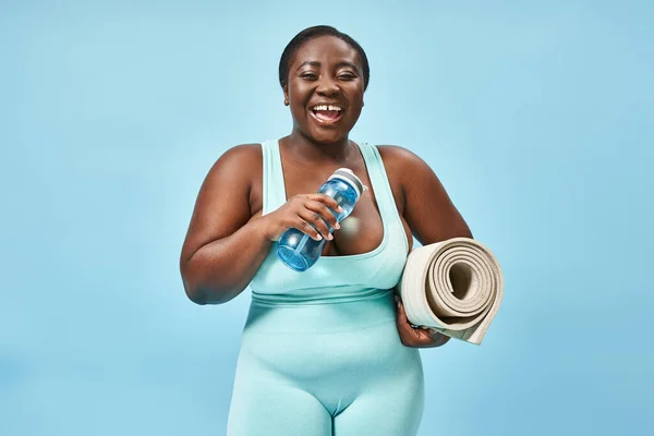 Smiling plus size african american woman standing with fitness mat and water bottle on blue — Stock Photo