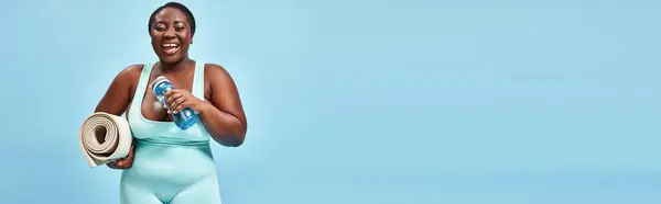 Веселый плюс размер африканский американец женщина стоит с фитнес мат и бутылку воды на синий, баннер — стоковое фото