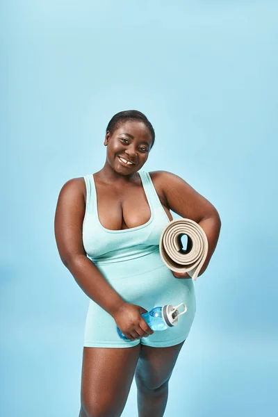 Alegre más tamaño afroamericano mujer de pie con tapete de fitness y botella de agua en azul - foto de stock