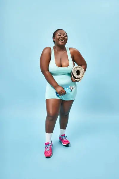 Sonriendo más tamaño mujer afroamericana de pie con tapete de fitness y botella de agua sobre fondo azul - foto de stock