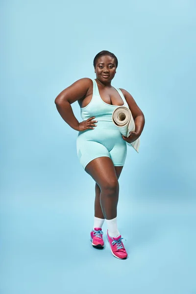 Sonriendo más tamaño mujer afroamericana de pie con alfombra de fitness sobre fondo azul, deporte - foto de stock