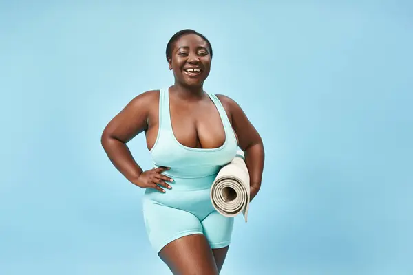 Smiling plus size african american woman standing with fitness mat on blue backdrop, body positive — Stock Photo
