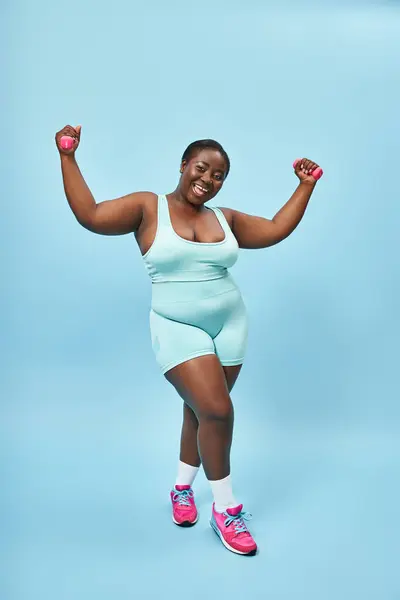 Feliz más mujer de tamaño en ropa deportiva azul con mancuernas disfrutando de su ejercicio sobre fondo a juego - foto de stock