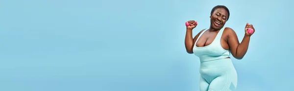 Happy plus size woman with dumbbells enjoying her work out on blue background, banner — Stock Photo