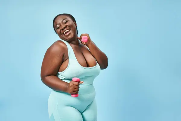 Happy plus size woman in blue active wear with dumbbells enjoying her work out on matching backdrop — Stock Photo
