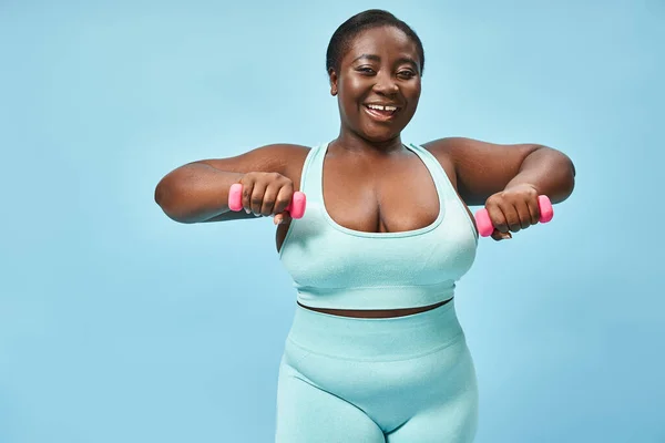 Positive plus size woman in blue active wear exercising with dumbbells on matching background — Stock Photo