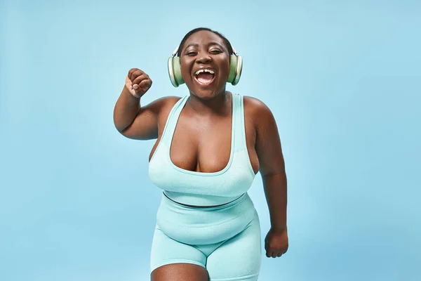 Cheerful plus size african american woman dancing in active wear with headphones on, blue background — Stock Photo