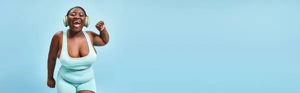 Счастливая плюс размер африканская американка танцует в активной одежде с наушниками на, баннер — стоковое фото