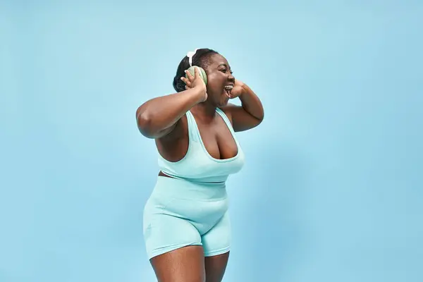 Happy plus size african american woman in active wear listening music in headphones, blue backdrop — Stock Photo