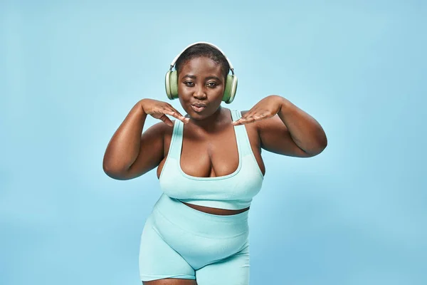 Jeune plus la taille femme afro-américaine en tenue active écouter de la musique dans les écouteurs, fond bleu — Photo de stock