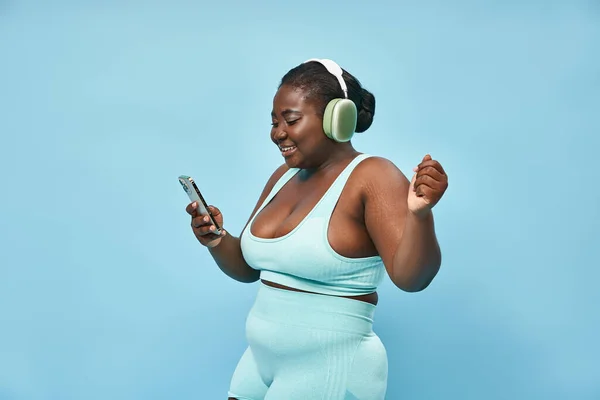 Plus size african american woman listening music in headphones and holding smartphone, blue backdrop — Stock Photo