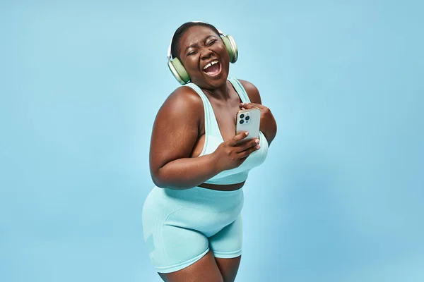 Plus size donna afroamericana ascoltando musica in cuffia e tenendo smartphone, blu — Foto stock