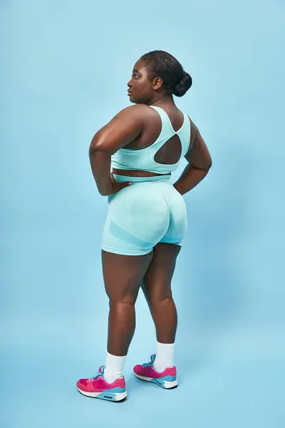 Más tamaño mujer afroamericana en ropa activa con estilo mirando hacia atrás y posando con las manos en las caderas - foto de stock