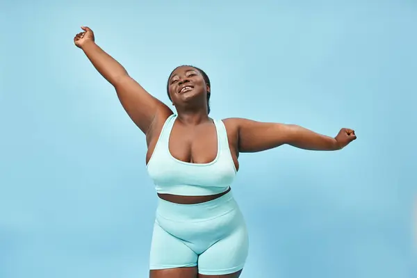 Glücklich plus size afrikanisch-amerikanische Frau in aktiver Kleidung, die sich freudig auf blauem Hintergrund dehnt — Stockfoto