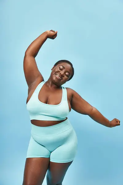 Gai plus la taille femme afro-américaine en tenue active s'étendant joyeusement sur fond bleu — Photo de stock