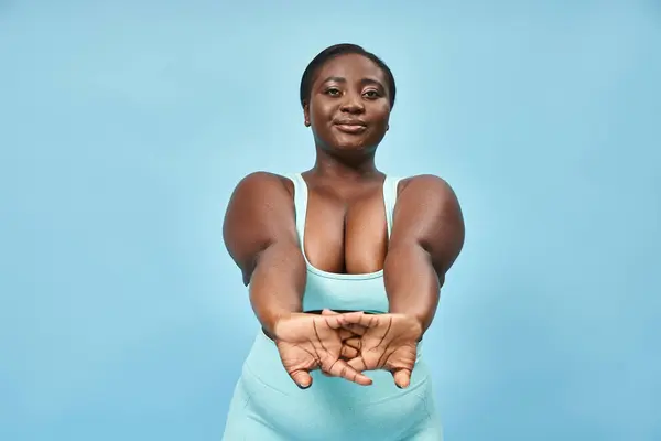 Feliz más tamaño mujer afroamericana en desgaste activo estiramiento sobre fondo azul, cuerpo positivo - foto de stock