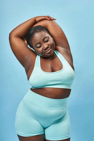 Pleased plus size african american woman in active wear stretching on blue background, body positive — Stock Photo