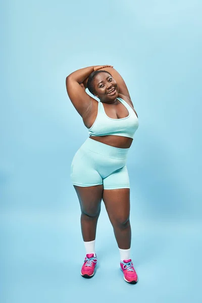 Complacido más tamaño mujer afroamericana en desgaste activo estiramiento sobre telón de fondo azul, cuerpo positivo - foto de stock