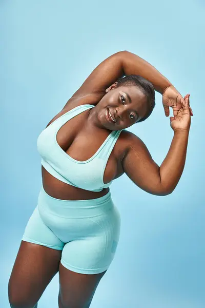 Весела афроамериканка плюс розмір в активному одязі, що тягнеться на синьому фоні, позитивне тіло — стокове фото