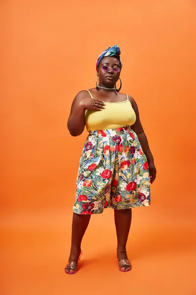 Elegante mujer afroamericana de talla grande con falda floral y pañuelo en la cabeza posando sobre fondo naranja - foto de stock