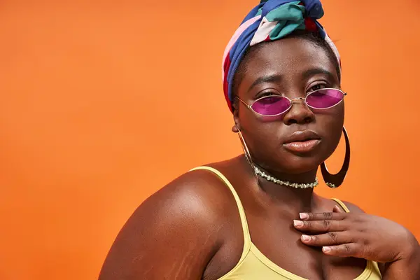 Fashion-forward et taille plus femme afro-américaine en foulard et lunettes de soleil tendance sur orange — Photo de stock