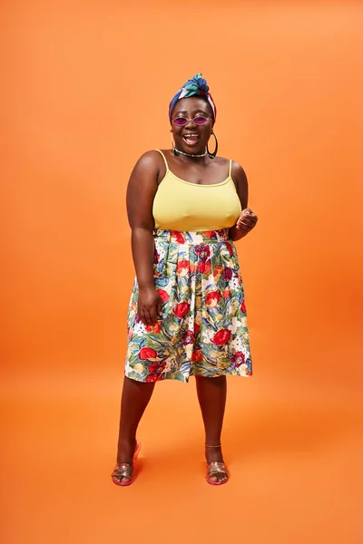 Excité afro-américain plus taille femme avec jupe florale et lunettes de soleil posant sur fond orange — Photo de stock