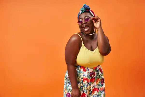 Счастливый африканский американец плюс размер женщина в цветочной юбке и солнцезащитные очки позируя на оранжевом фоне — стоковое фото
